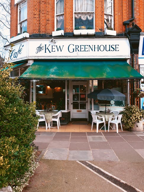 The image shows the front exterior of a café called 'The Kew Greenhouse,' situated in a red-brick building with large upstairs windows partially covered by lace curtains. Below the signage, there is a green awning shading the entrance. In front of the café, on the paved pavement, there are two outdoor seating areas with white plastic chairs and round tables; one table has a dark-colored umbrella providing shade. Potted plants with greenery and flowering bushes are positioned on either side of the entrance, enhancing the curb appeal. The scene is lit with natural daylight, creating a welcoming atmosphere consistent with a typical establishment offering outdoor service or al fresco dining. This setting may relate to alternative disposal or in-situ clearing efforts, as captured in a context where rubbish removal services could be involved, supporting private waste management solutions for local businesses like this café.
