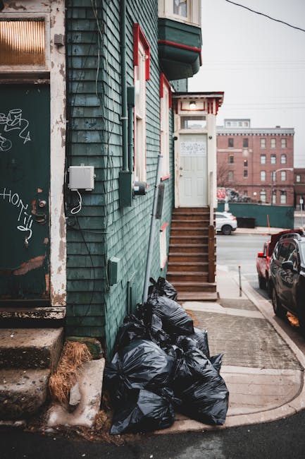 A vertical view of a building's exterior featuring dark green wooden cladding with a textured, weathered finish. On the left side, there is a partially visible door with graffiti and rusted metal elements, alongside a small white electrical box and a grey cable. In the center, a set of wooden stairs with natural wood finish and red-painted risers leads up to a white entrance door, which has a small window and a decorative awning above it. At the base of the stairs, several tightly packed black plastic rubbish bags are stacked on the pavement, with some foliage and debris nearby. To the right, parked cars are visible along a street with modern brick and concrete buildings in the background, under an overcast sky. The scene depicts an area where private rubbish collection might be carried out, with rubbish bags awaiting removal, fitting with the context of independent waste management services.