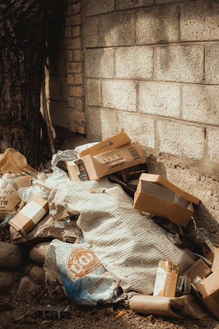 A pile of discarded cardboard boxes and recyclable packaging materials, some partially flattened and others still intact, resting against a weathered brick wall and rough concrete blocks. The boxes vary in size and are primarily brown with printed labels and barcodes, some torn or crumpled. Accompanying the cardboard are a few plastic bags containing additional waste, all situated on uneven ground composed of small rocks and dirt. The scene appears to be an outdoor informal rubbish collection area, possibly on a driveway or back alley. The lighting is natural, casting soft shadows around the debris, and there are no visible tools or equipment. The presence of these waste materials suggests a local, independent collection or on-site clearance scenario, typical of a private waste handling setup, aligning with the services of companies like Rubbish Clearance Richmond upon Thames. This image illustrates the process of waste stacking before removal or disposal in an environmental context supporting rubbish removal services or alternative waste handling solutions.