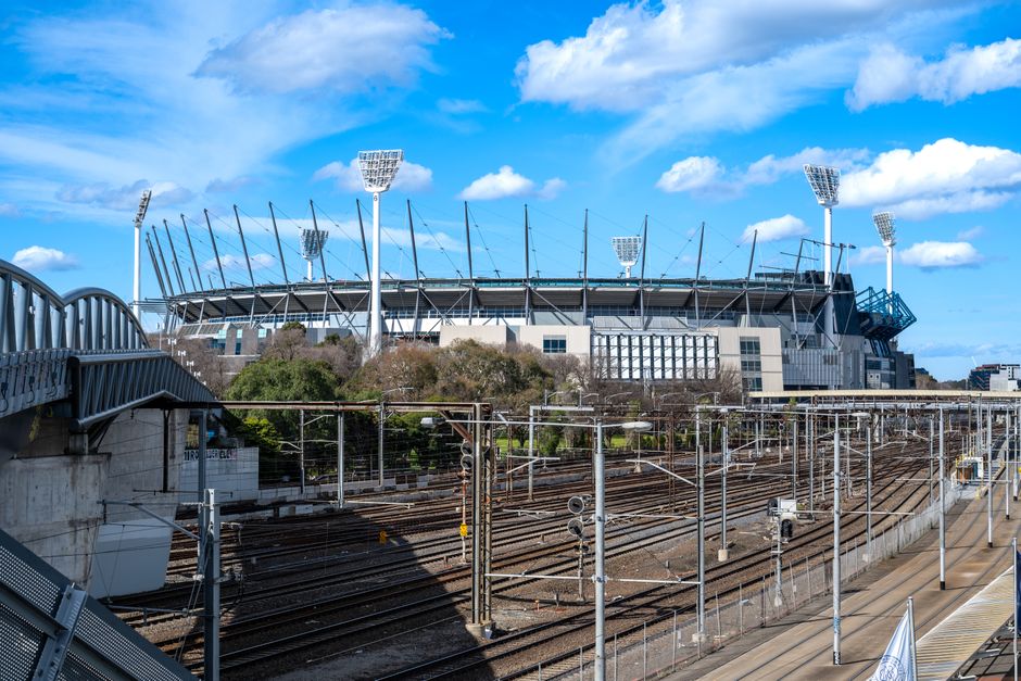 The image shows a large modern sports stadium with a curved architectural design, surrounded by tall stadium floodlights with rectangular light panels extending above the structure. In the foreground, there are multiple railway tracks with overhead electrical wiring supported by numerous metal poles and gantries, running parallel to a concrete platform on the right side. To the left, a metal pedestrian bridge with a curved railing spans across the railway lines, connecting to the stadium area. The sky is bright and partly cloudy, casting natural daylight over the scene, highlighting the metallic textures of the stadium’s exterior and the railway infrastructure. The environment appears to be an urban setting, with no visible natural vegetation in the immediate vicinity of the train tracks and stadium, emphasizing an area dedicated for transportation and large-scale sporting events. The scene suggests a context related to the infrastructure of the stadium, possibly indicating the importance of efficient access and logistics, which aligns with services like independent rubbish collection or waste management support for large public venues, as provided by companies such as rubbishclearancerichmonduponthames.com.