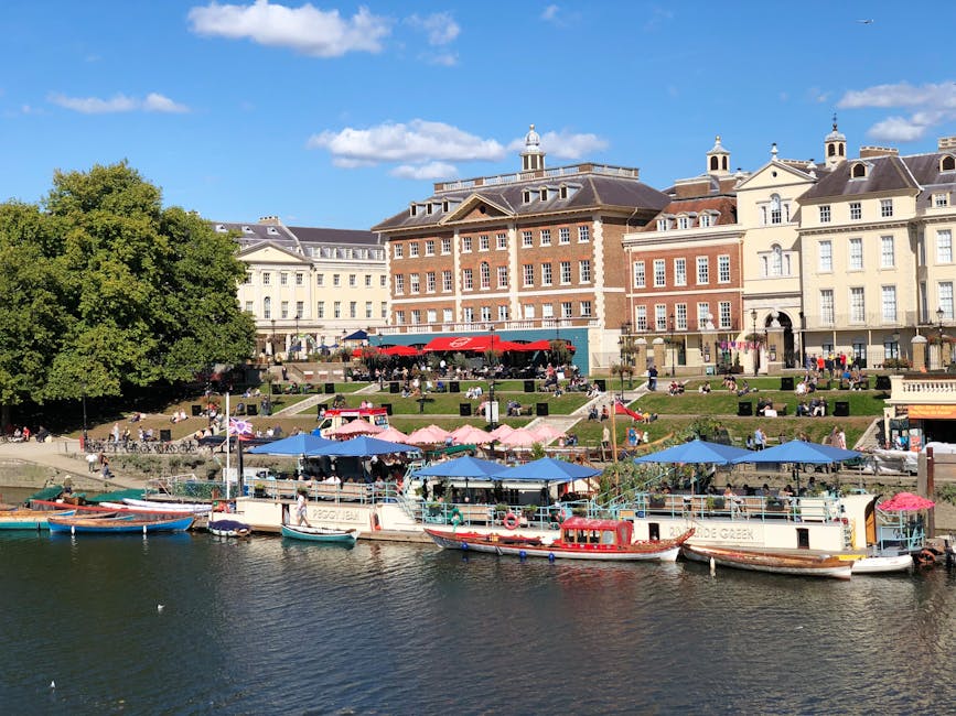 A daytime view of a riverside area with several small boats docked along the water's edge, featuring bright blue, red, and wooden finishes, moored next to a promenade adorned with multiple outdoor seating areas shaded by pink and blue umbrellas. In the background, there is a large, multi-story building with a mix of brick and light-colored facades, decorative architectural elements, and small tower-like structures on the roof, set against a partly cloudy sky. The scene includes a gently sloping grassy area with people relaxing and walking, creating a lively atmosphere typical of urban riverside leisure spaces. This outdoor setting, with its varied textures and colors, may relate to independent waste management practices where rubbish removal services could be used to maintain cleanliness in such public areas, reflecting alternatives to traditional municipal collection in busy, well-frequented locations near Richmond station.